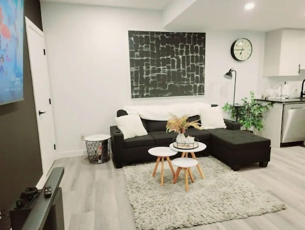 Un salón moderno con un sofá negro, un tapete blanco y una mesa de centro con tres taburetes. Un cuadro abstracto de gran tamaño cuelga de la pared, y un reloj se ve al fondo.
