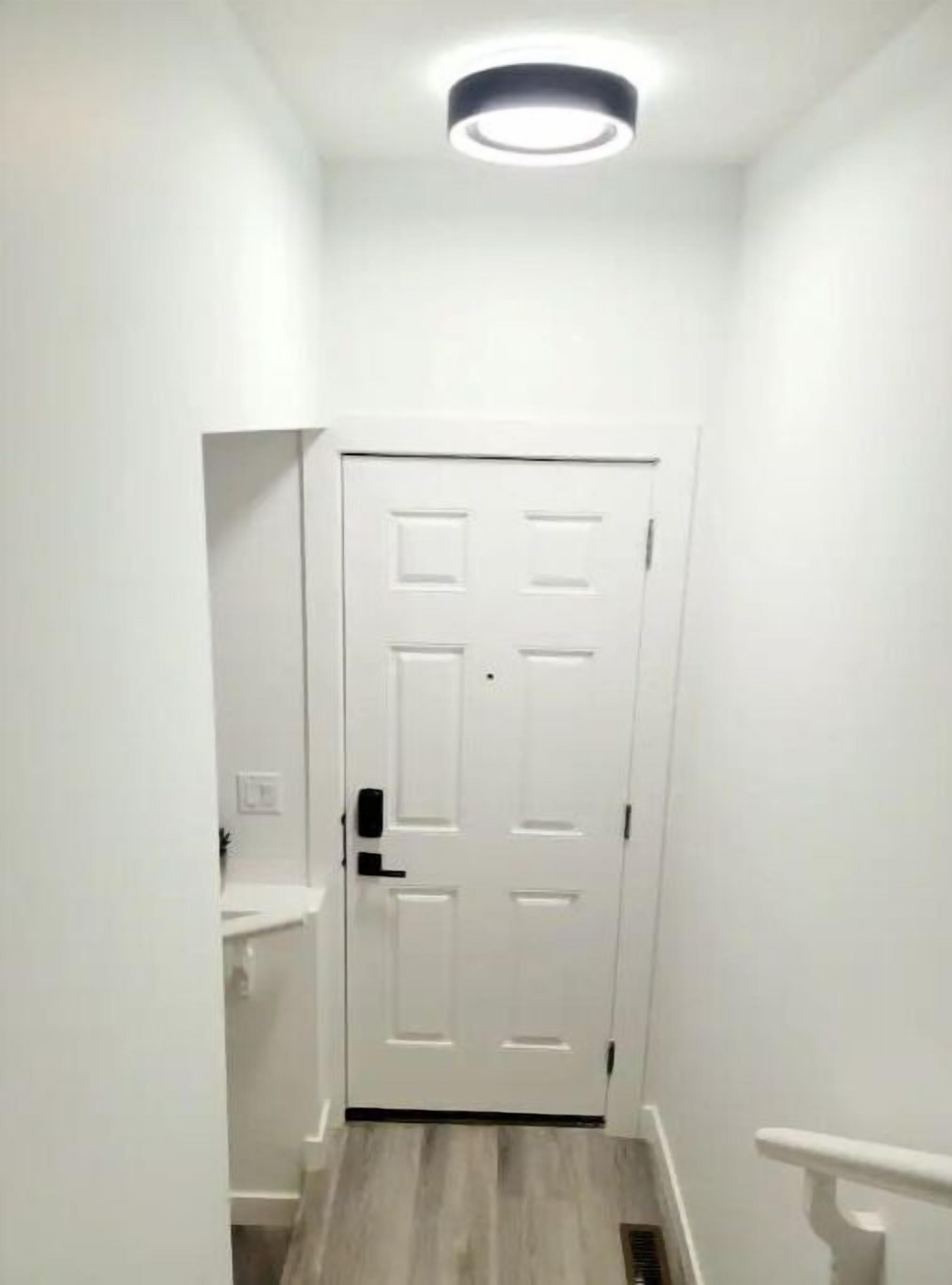 Un pasillo estrecho con una puerta blanca, una lámpara arriba y un estante pequeño a la izquierda.
