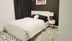A modern bedroom with a white bed, black and white pillows, a nightstand with a lamp, and a geometric patterned rug. - Luxury Living Basement suite (Calgary)