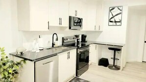 A modern kitchen with white cabinets, a black countertop, and a small dining area with a stool. - Luxury Living Basement suite (Calgary)