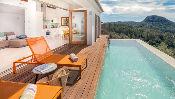 A modern outdoor pool area with wooden decking, lounge chairs, and a view of a mountainous landscape.