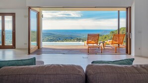 A room with a large sliding glass door leading to a balcony with a view of a beach and mountains. There are two wooden chairs on the balcony.