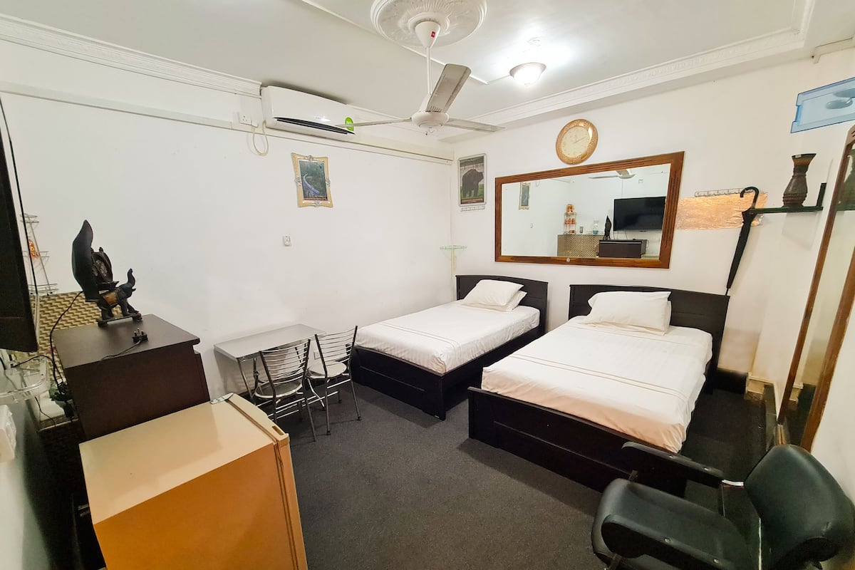Ein Hotelzimmer mit zwei Einzelbetten, einem Schreibtisch mit Stuhl, einem kleinen Fernseher, einem Deckenventilator und einem Spiegel.