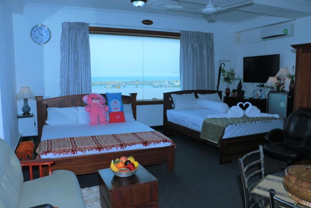 A hotel room with two beds, a sofa, a dining table with fruit, a TV, and a view of the sea.