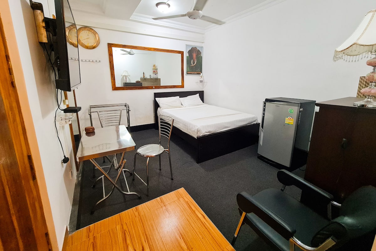 Ein kleines Hotelzimmer mit einem Bett, einem Schreibtisch, einem Sessel, einem Fernseher und einem Deckenventilator.