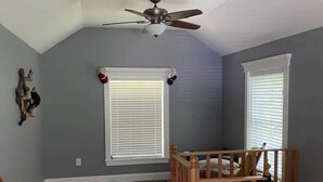 Ein Zimmer mit schrägen Wänden, Deckenventilator, zwei Fenster mit Jalousien, eine Holztreppe und ein kleiner Teppich auf dem Boden.