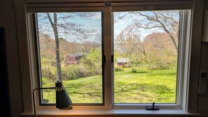 Una habitación con un ventanal grande que da a un espacio verde con árboles y un edificio pequeño. Una lámpara de escritorio está sobre el alféizar.