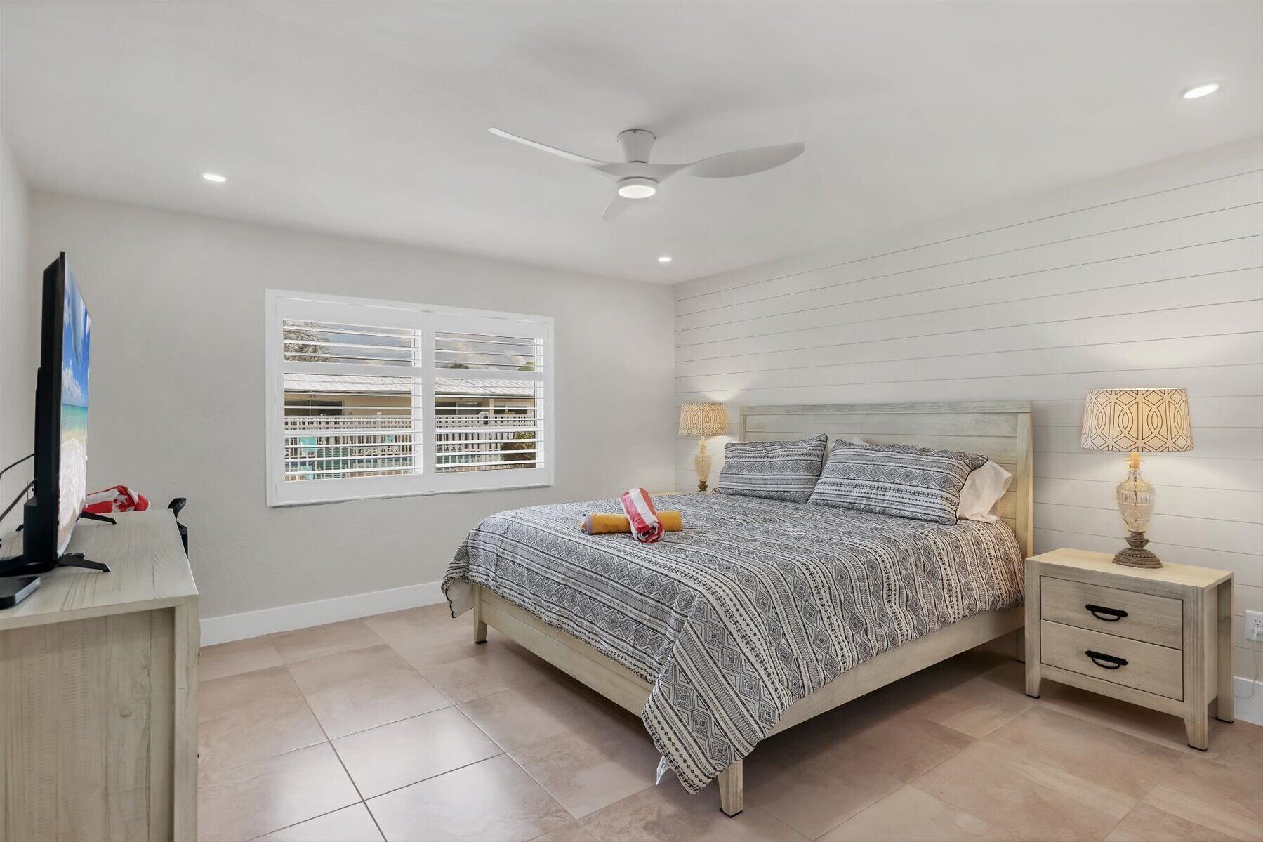 A bedroom with a bed, bedside tables, a ceiling fan, a window with blinds, and a TV on a stand.
