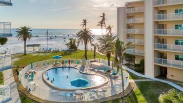 A pool area with a circular design, surrounded by lounge chairs and umbrellas, overlooking a beach with palm trees and a building with balconies.