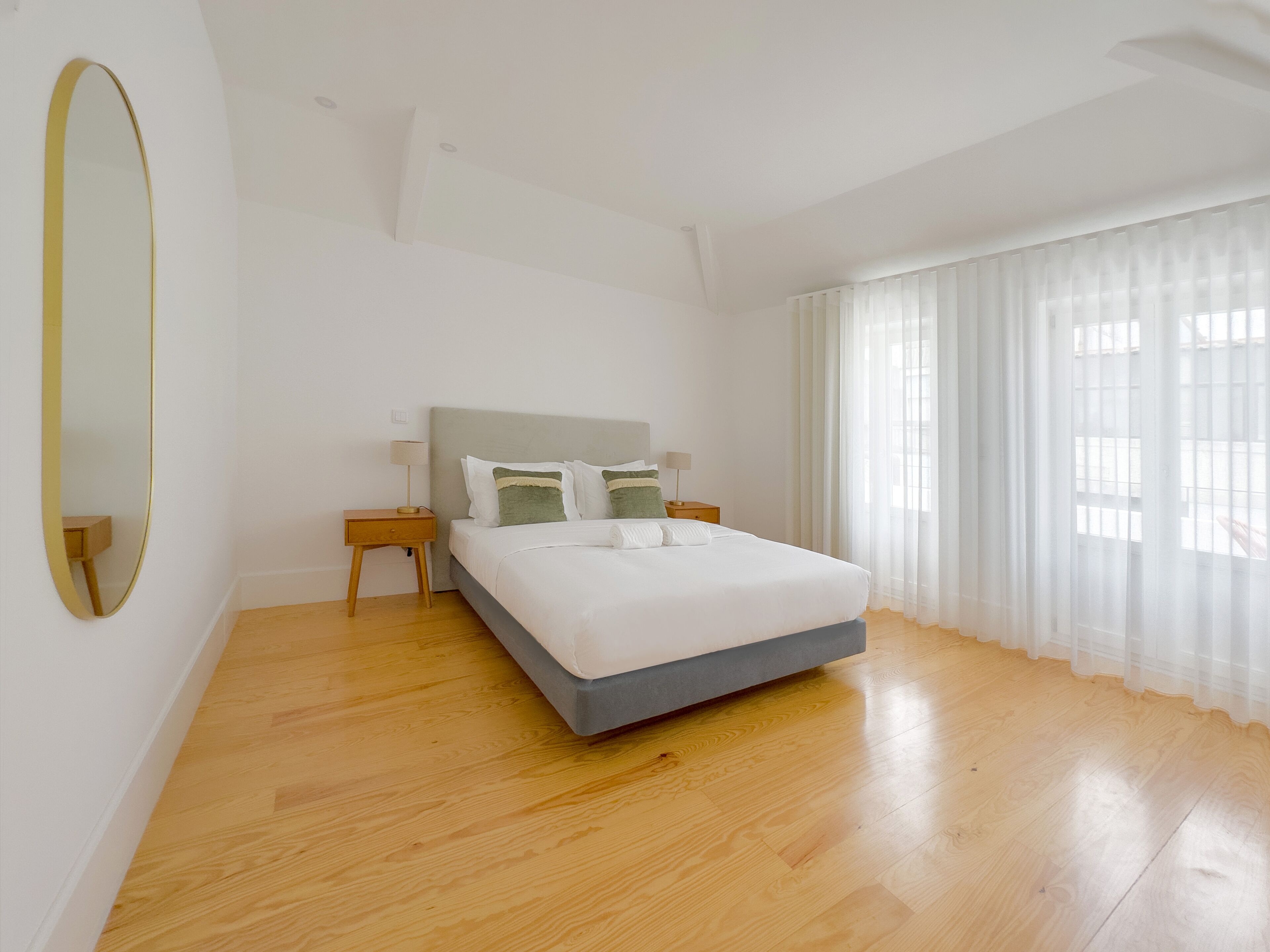 Een slaapkamer met een bed, nachtkastje, spiegel en een raam met gordijnen.