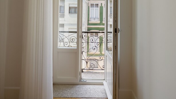 Una habitación con piso de madera, paredes blancas y una puerta que da a un balcón con vista a edificios.