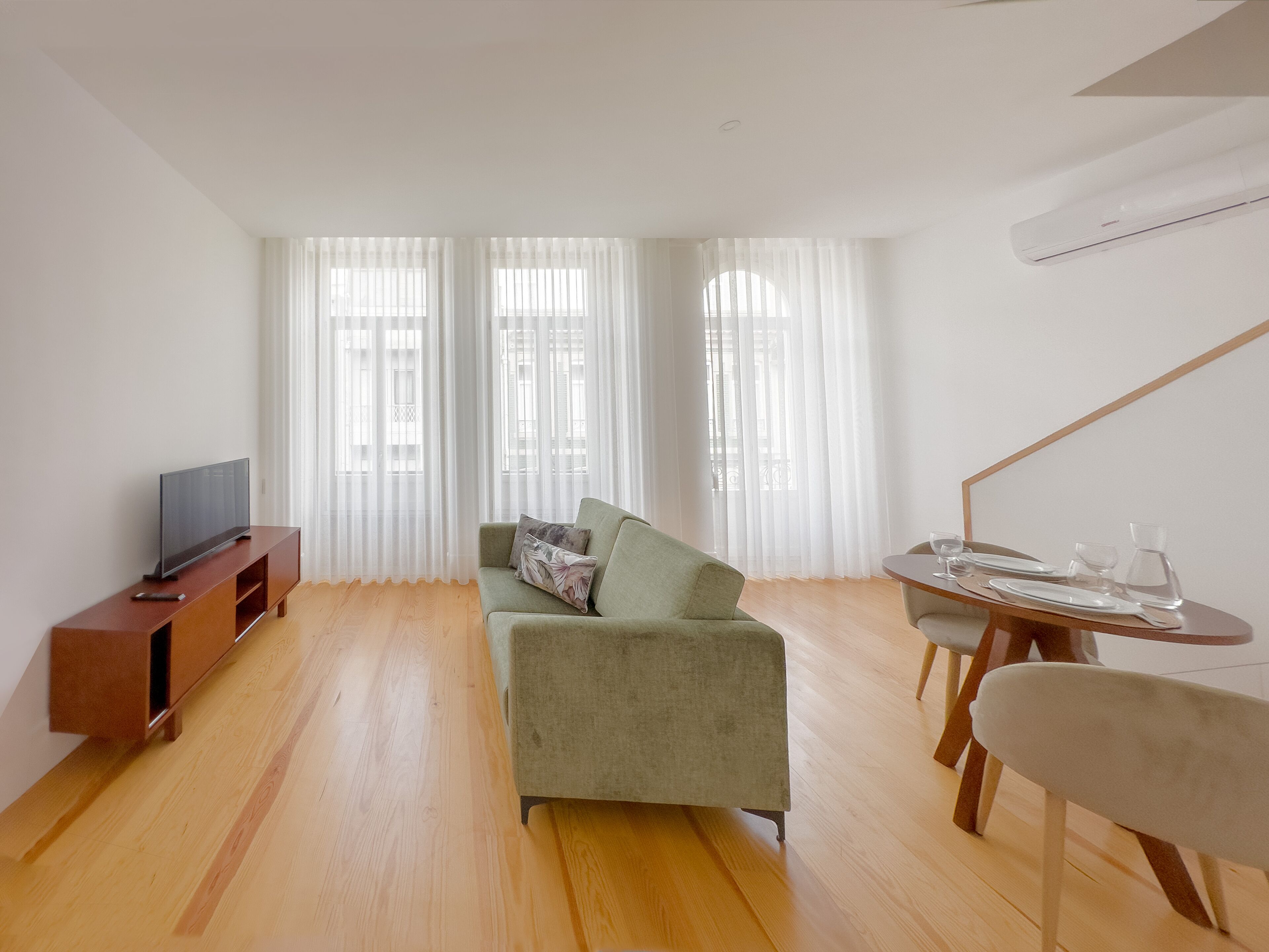 A modern living room with a flat-screen TV, a green sofa, a dining table with chairs, and large windows with sheer curtains.