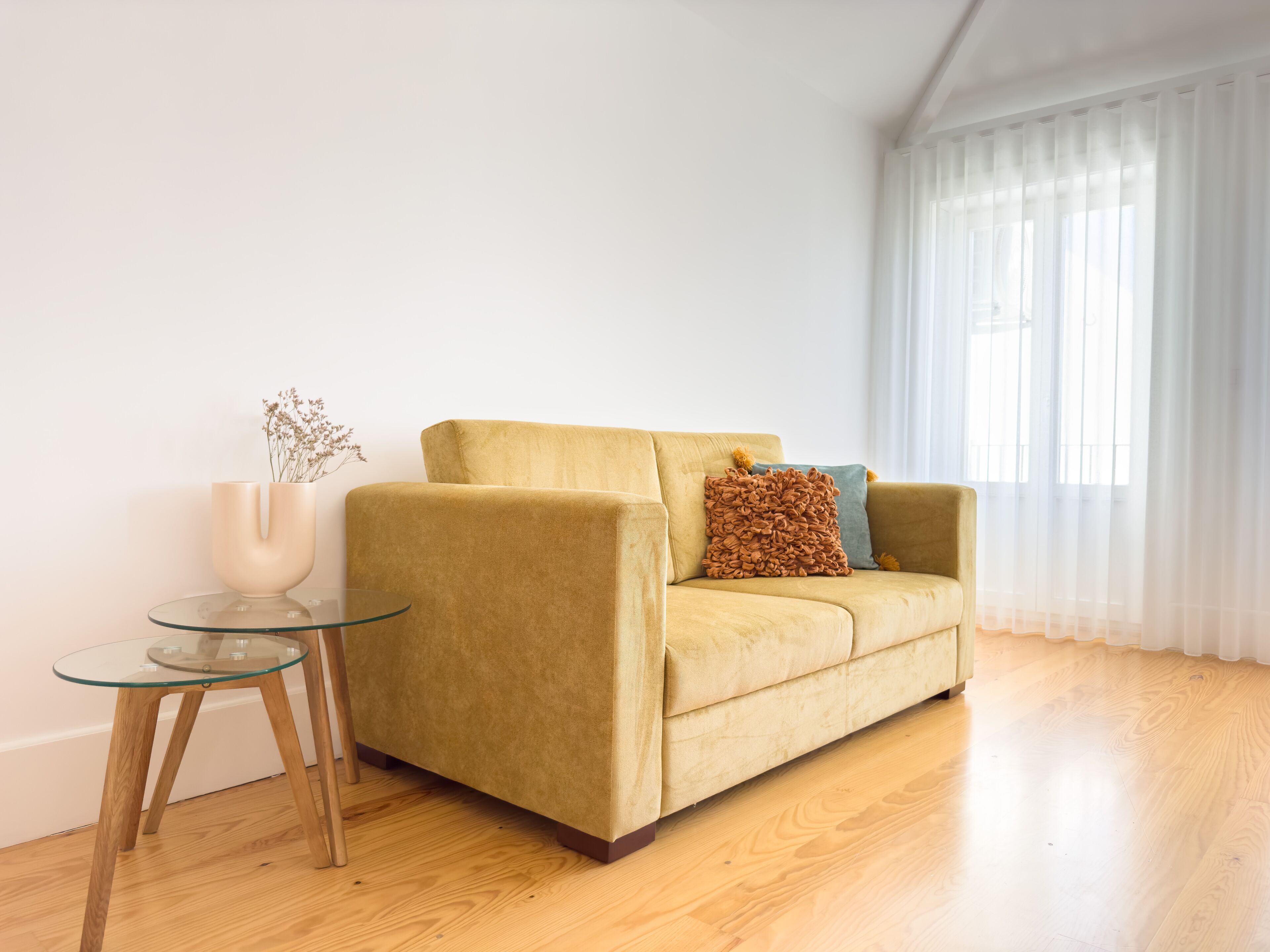Ein gelbes Sofa mit einem braunen und blauen Kissen, ein Glastisch mit einer Blumenvase und ein großes Fenster mit transparenten Vorhängen.