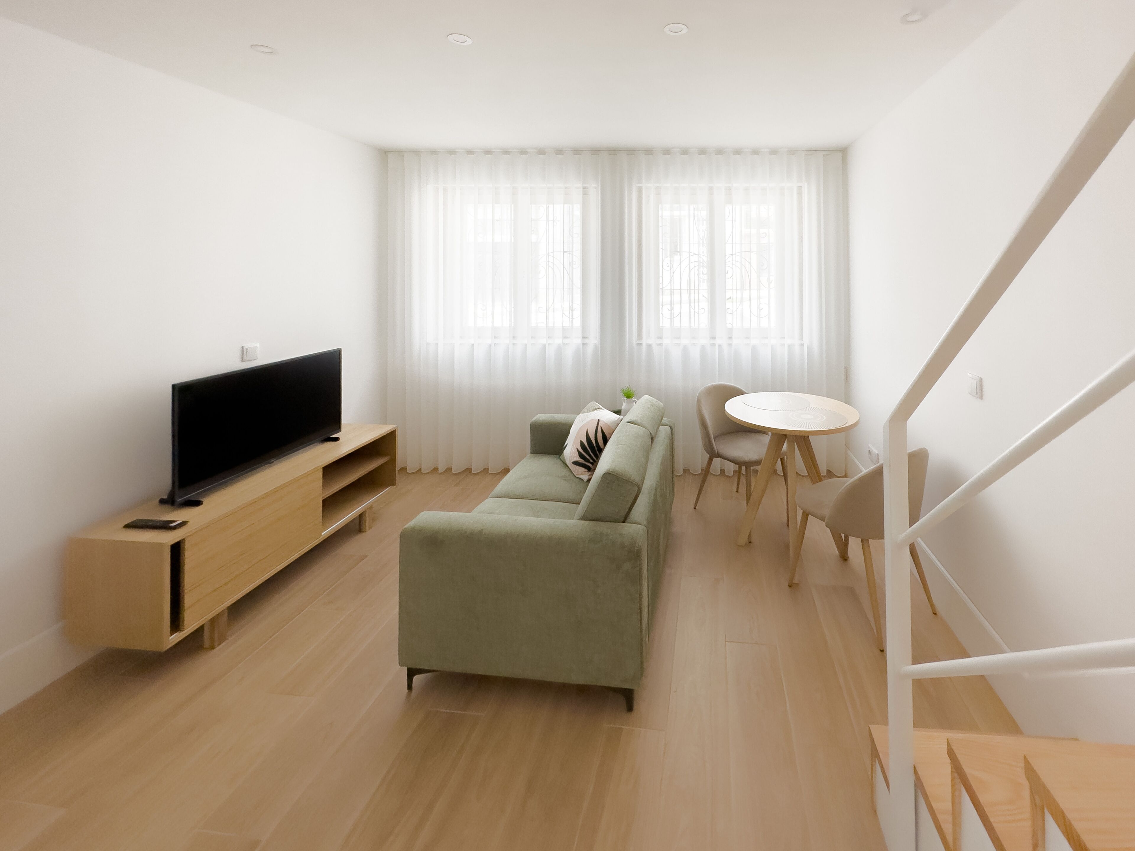 Una sala de estar moderna con un televisor de pantalla plana sobre un soporte de madera, un sofá verde, una pequeña mesa redonda y una escalera.