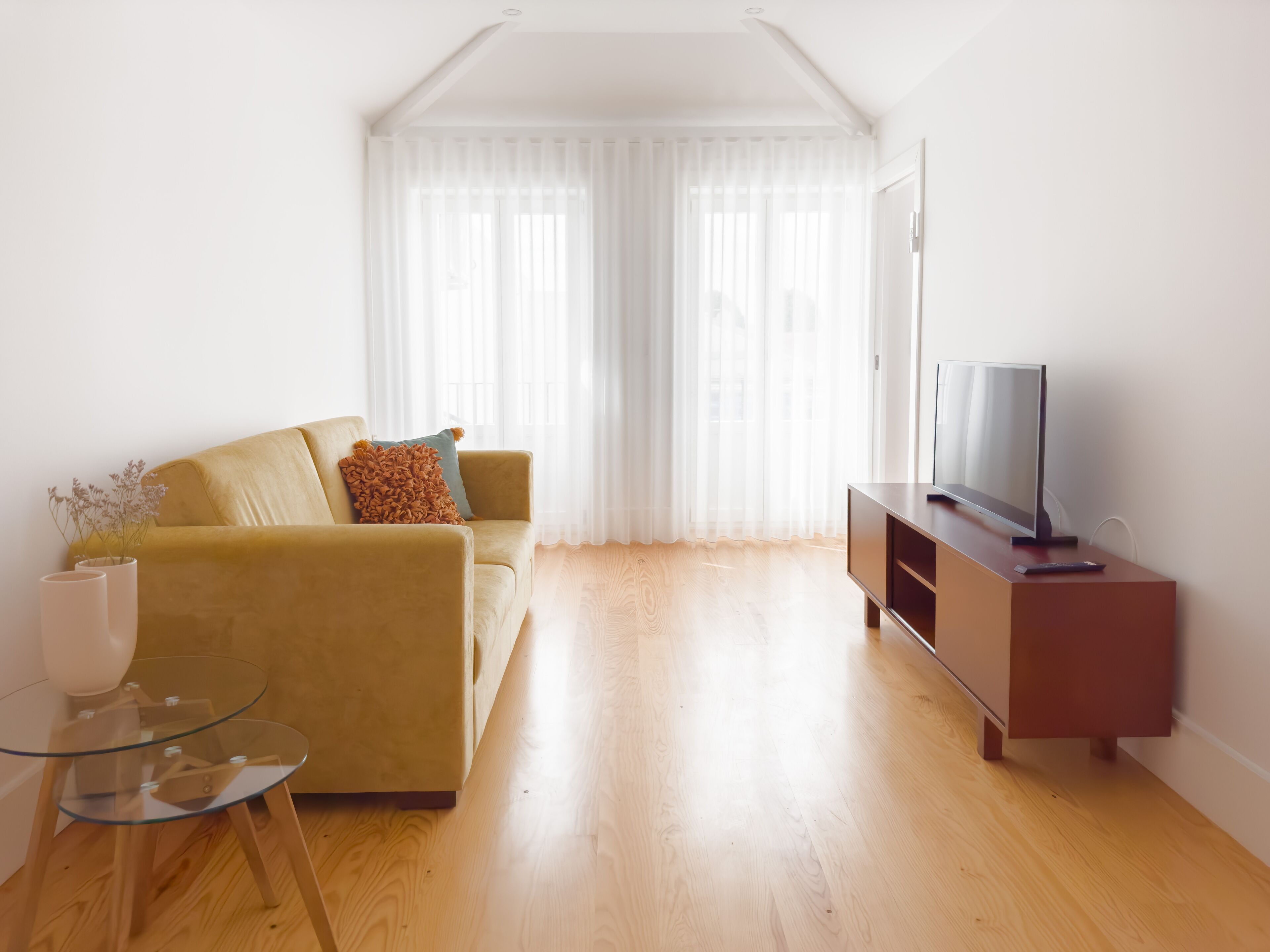 Una sala de estar con sofá, una mesa de centro de vidrio, un televisor sobre un mueble de madera y cortinas translúcidas.