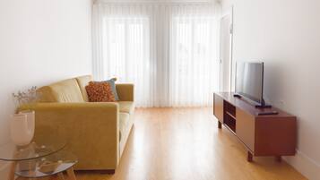 Una sala de estar con sofá, una mesa de centro de vidrio, un televisor sobre un mueble de madera y cortinas translúcidas.