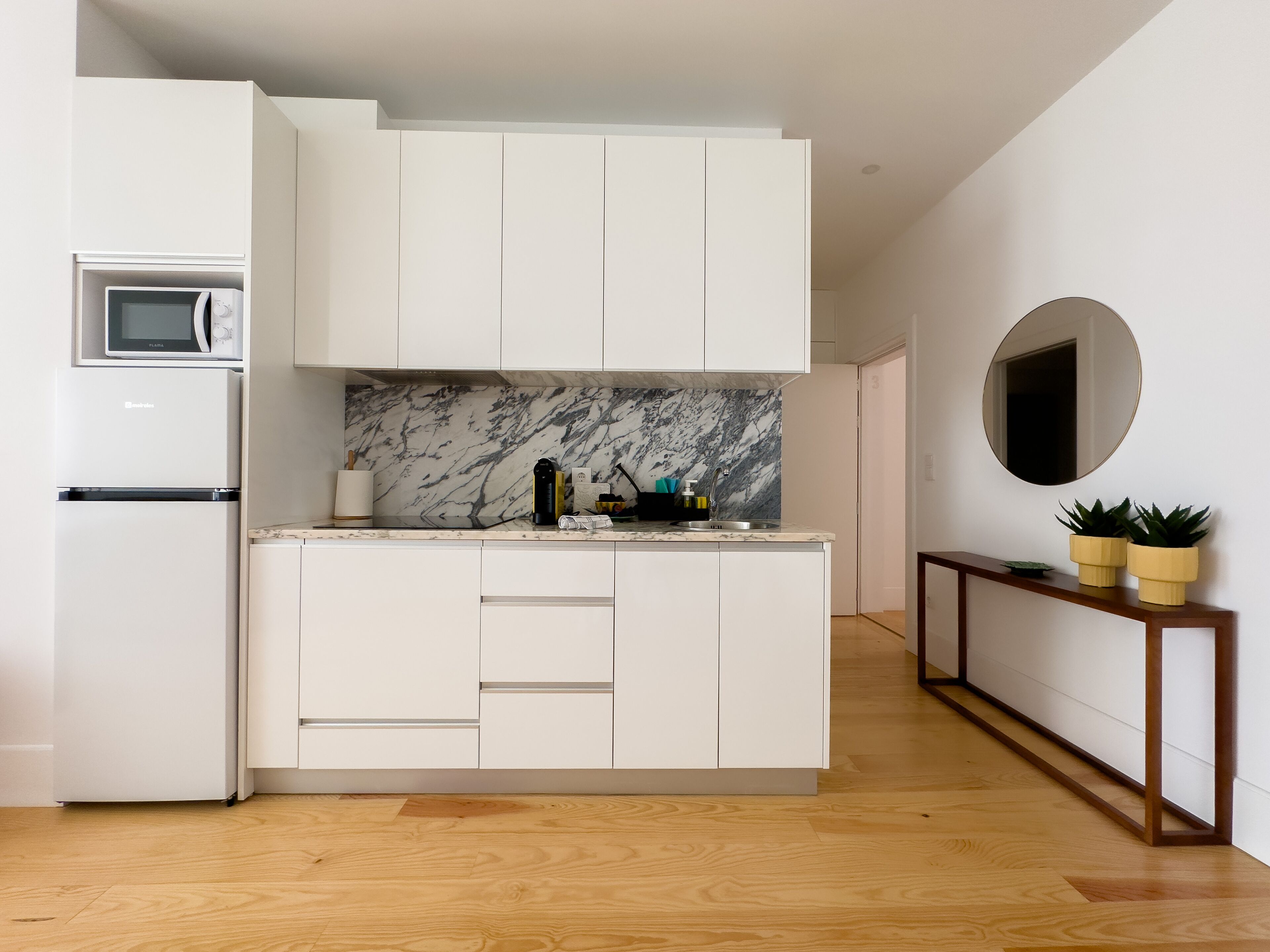 Una cocina moderna con armarios blancos, salpicadero de mármol, microondas integrado y una consola de madera con plantas en macetas.