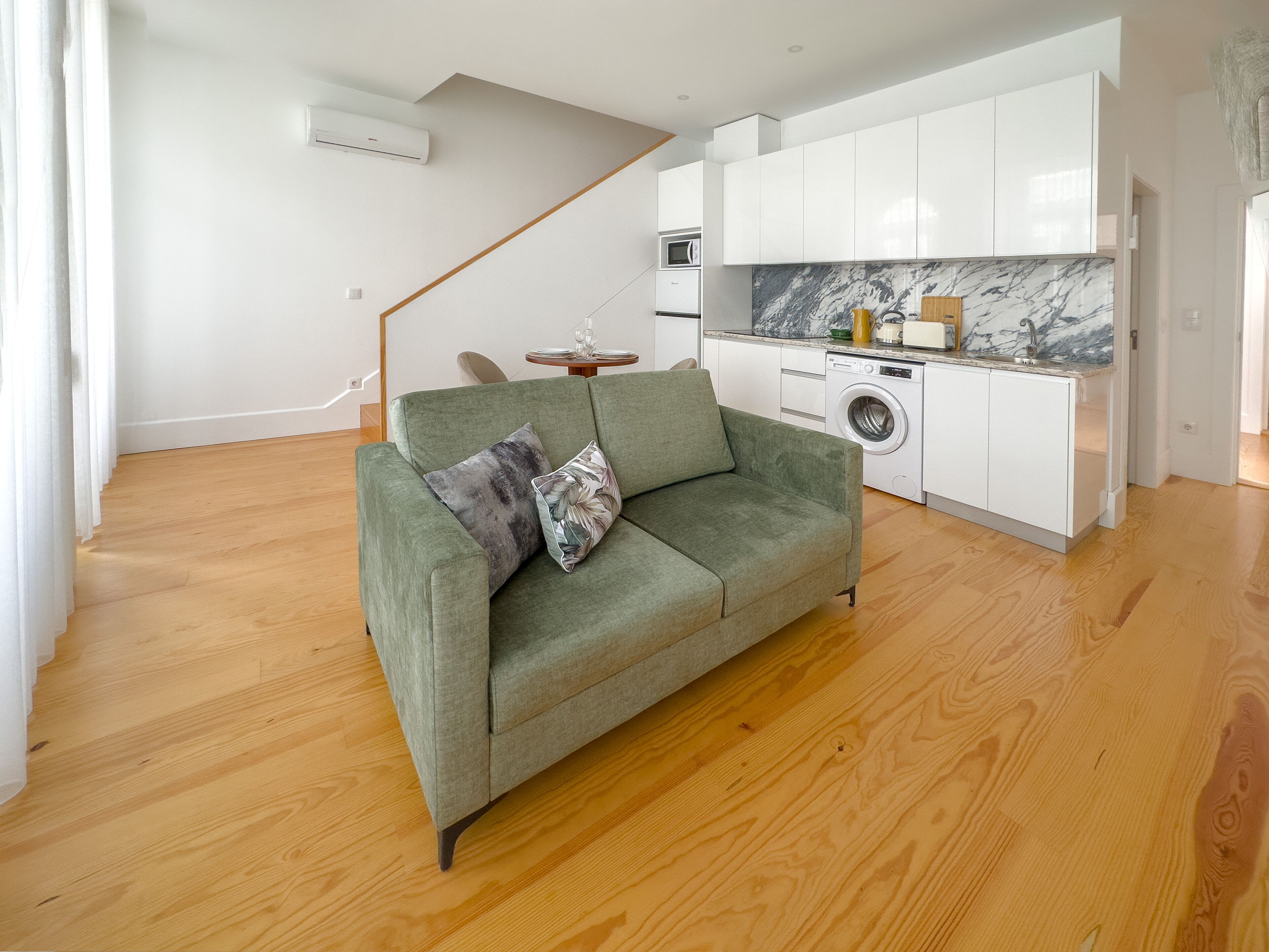 A modern apartment with a green sofa, a washing machine, and a kitchen with white cabinets and a marble backsplash.