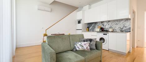 A modern living room with a green sofa, a marble backsplash in the kitchen, and wooden flooring.