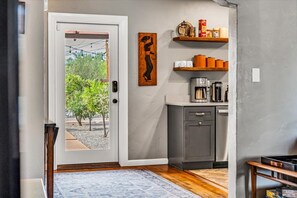 Cozinha com cafeteira, prateleiras com utensílios de cozinha e porta que dá para área externa com árvores.