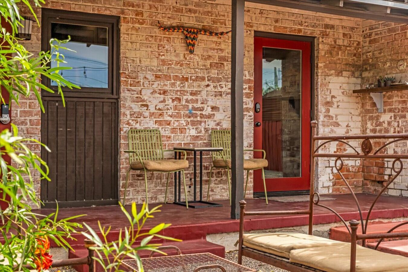 A brick building with a red door, two green chairs, a small table, and a metal railing.