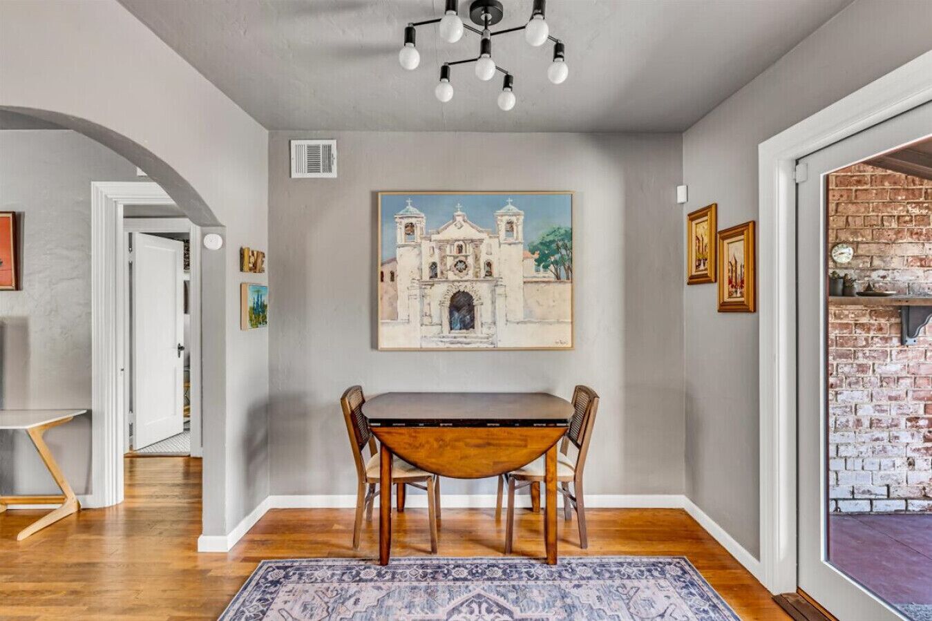 A dining area with a wooden table and chairs, a painting of a building on the wall, and a brick wall on the right.