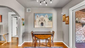 A dining area with a wooden table and chairs, a painting of a building on the wall, and a brick wall on the right.