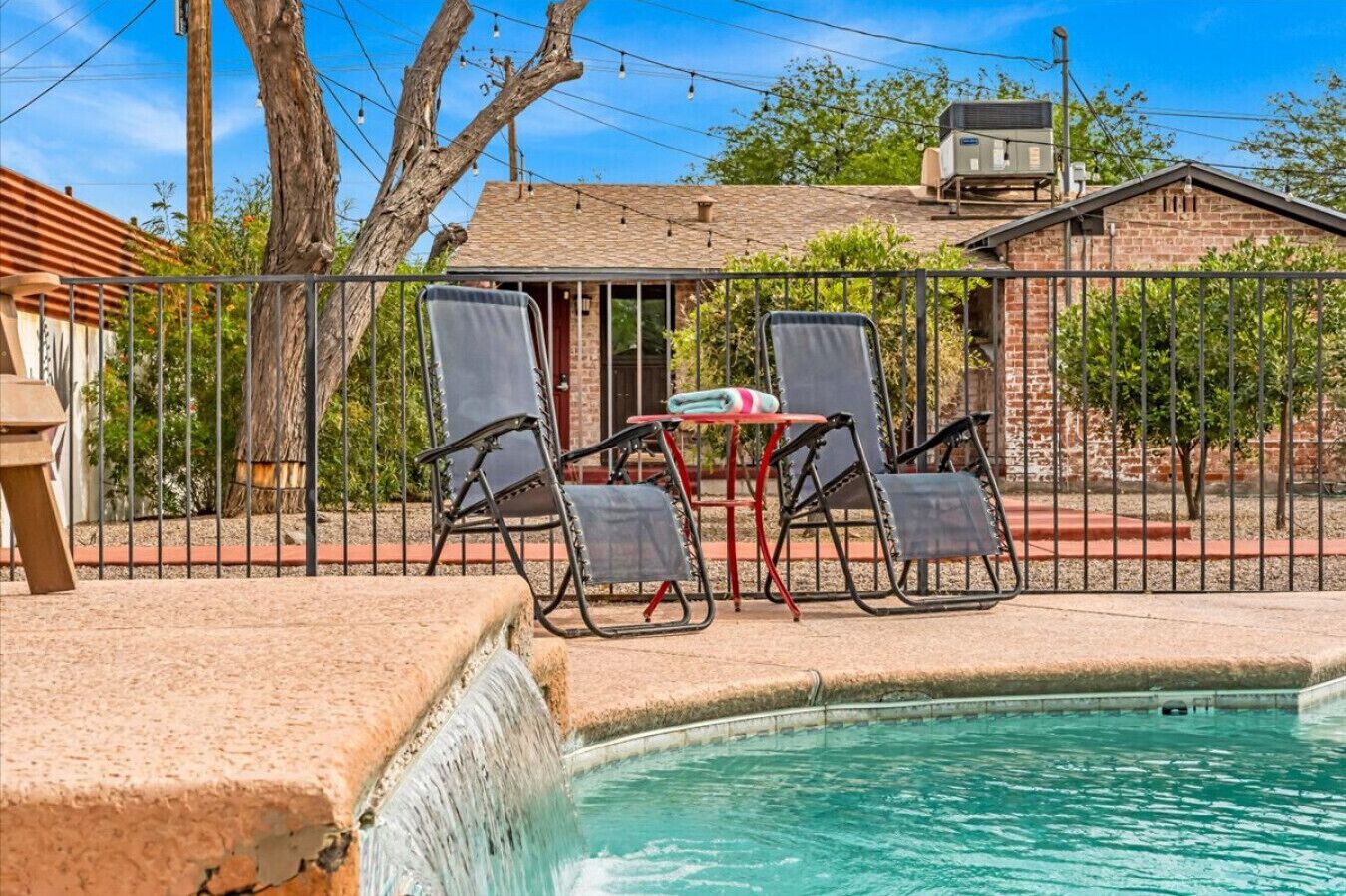 A swimming pool with a waterfall feature, surrounded by a fence, with two lounge chairs and a table with a towel.