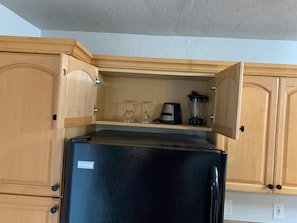Une cuisine avec des meubles de rangement en bois, un réfrigérateur noir et un plan de travail sur lequel se trouvent un mixeur et des verres.