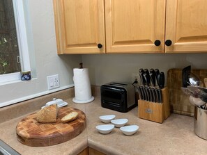 Une cuisine avec des meubles en bois, un grille-pain, une planche à découper avec du pain, un bloc à couteaux et une fenêtre donnant sur un beau paysage.