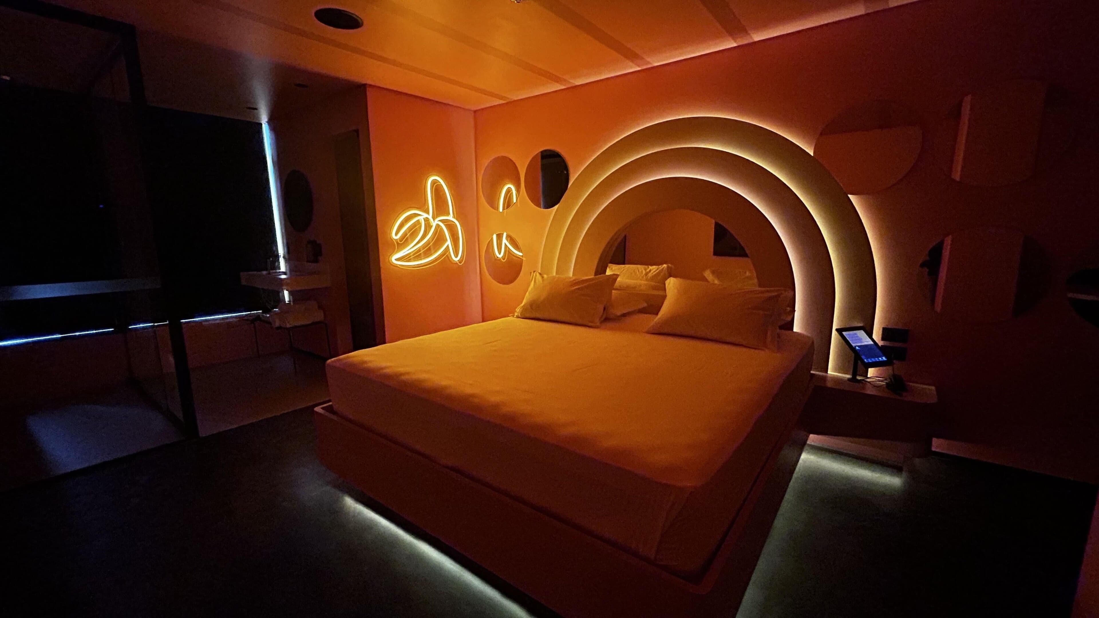 Ein modernes Schlafzimmer mit einem großen Bett, einem Nachttisch mit einem digitalen Gerät und einem Neon-Schild an der Wand.