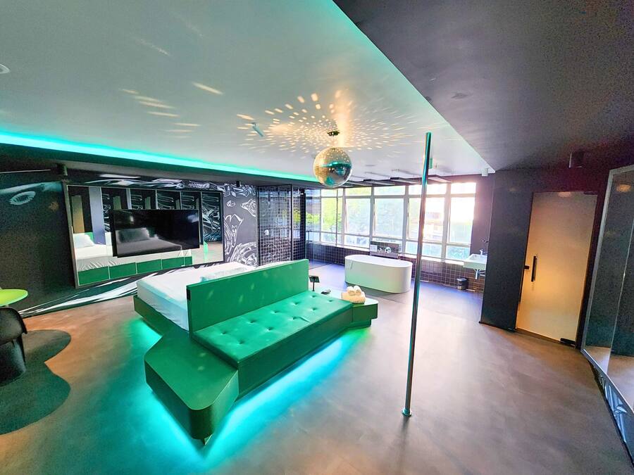 Ein modernes Hotelzimmer mit einem grünen Sofa, einem Flachbildfernseher, einem großen Fenster und einer Badewanne.