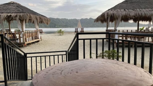 Una terraza frente al mar con sombrillas de paja y muebles de madera, con vista a una playa arenosa y un mar tranquilo.