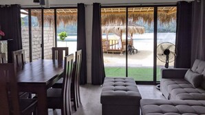 Una sala moderna con comedor, puertas correderas de cristal grandes que dan a vista de playa, una sombrilla de paja y un ventilador.