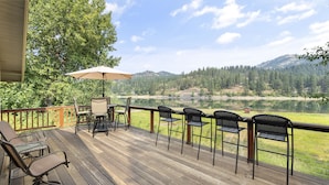 Eine Holzterrasse mit Gartenmöbeln, darunter ein Tisch und Stühle, mit Blick auf einen See und bewaldete Hügel.