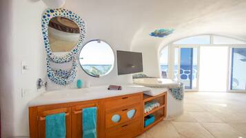 A bathroom with a large vanity, a round mirror, a television, and a view of the sea through large windows.