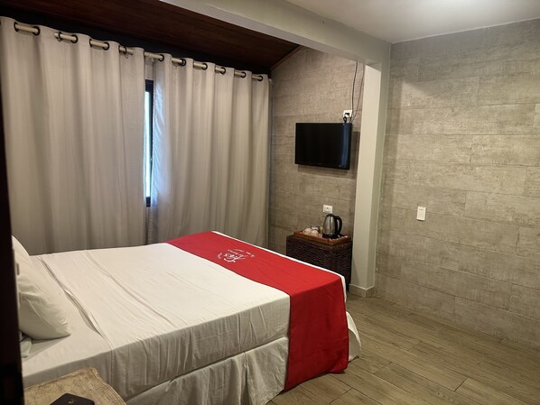 Quarto com cama, televisão na parede e uma mesinha com chaleira e chávenas.