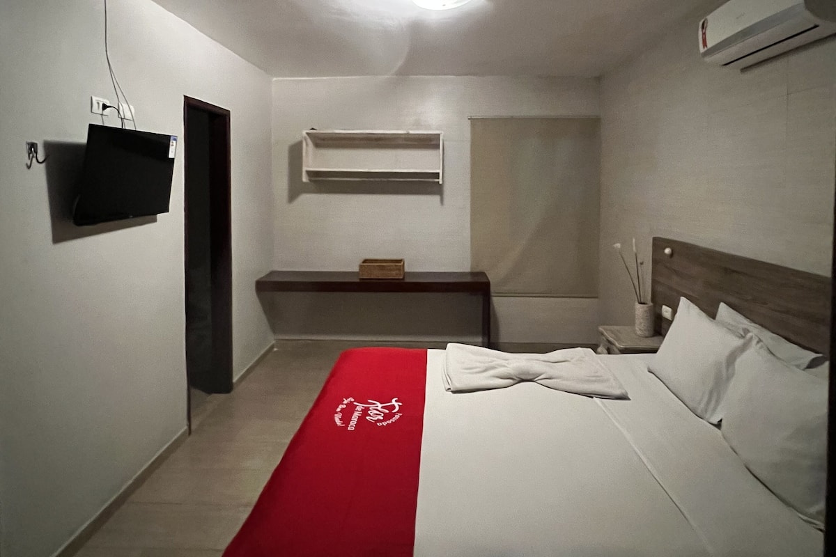 Hotellihuone, jossa on sänky, seinälle asennettu televisio, yöpöytä lampun kanssa ja seinälle asennettu ilmastointilaite.