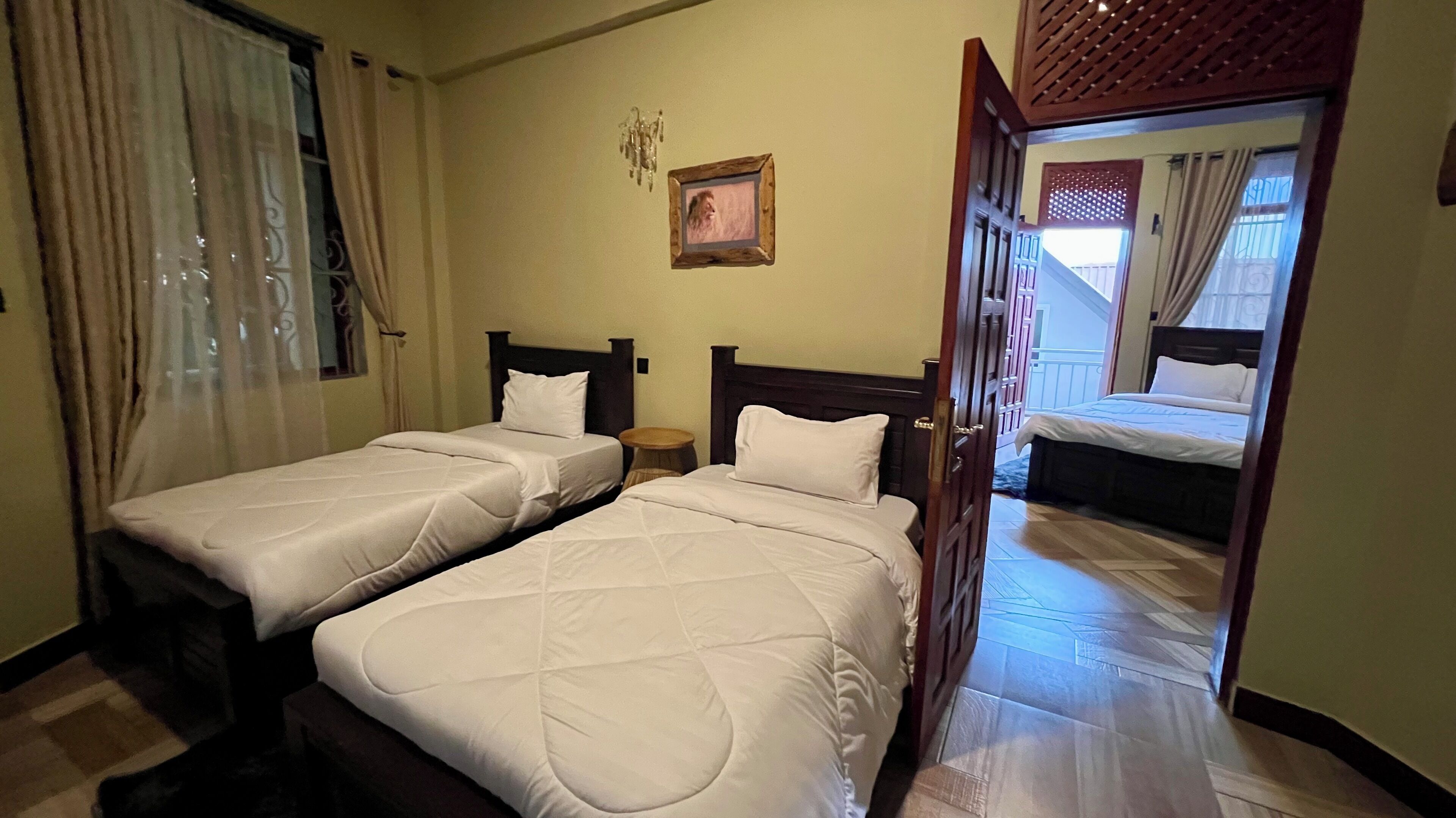 A hotel room with two single beds, a small round table, a wall-mounted picture, and a view of another room through an open door.