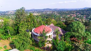 A circular building with a red roof surrounded by dense greenery, overlooking a hilly landscape with scattered houses.