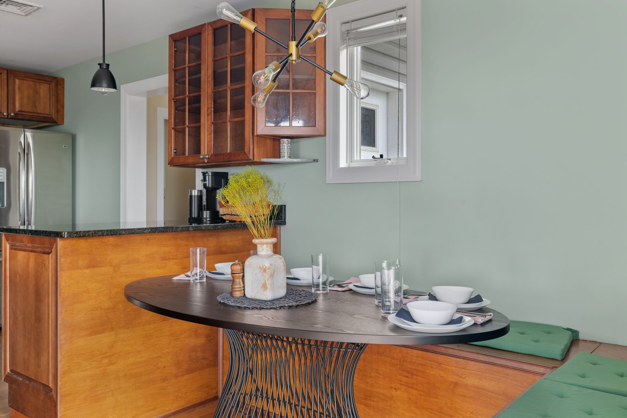 Une cuisine avec des meubles de rangement en bois, un îlot central doté d’un socle en osier, un luminaire suspendu et une fenêtre avec des stores.