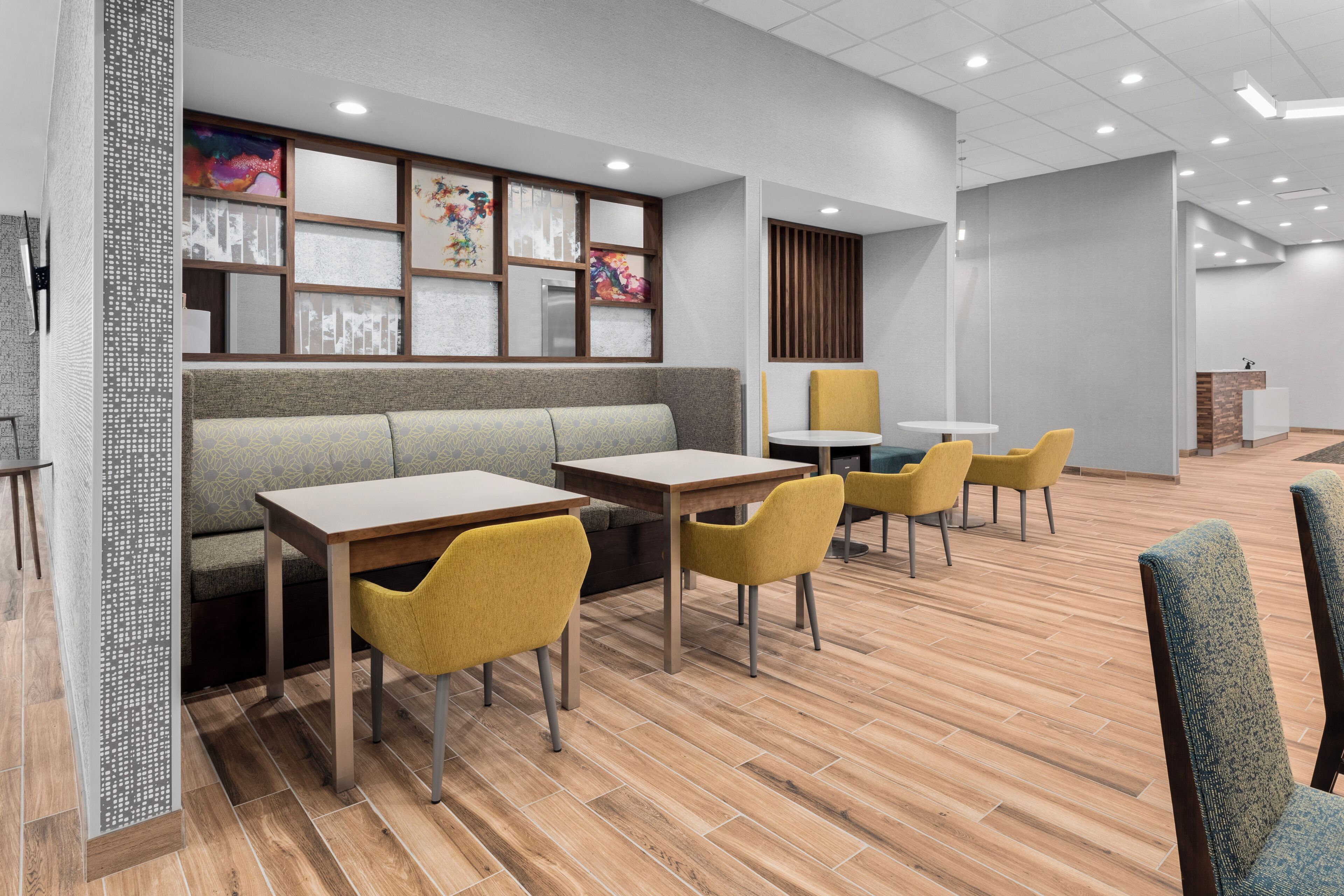 A modern restaurant with a wooden floor, yellow chairs, a grey sofa, and a wall with framed artwork.