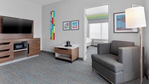 Une chambre d’hôtel moderne équipée d’un téléviseur à écran plat, d’un meuble de rangement en bois, d’un canapé gris et d’un lampadaire.