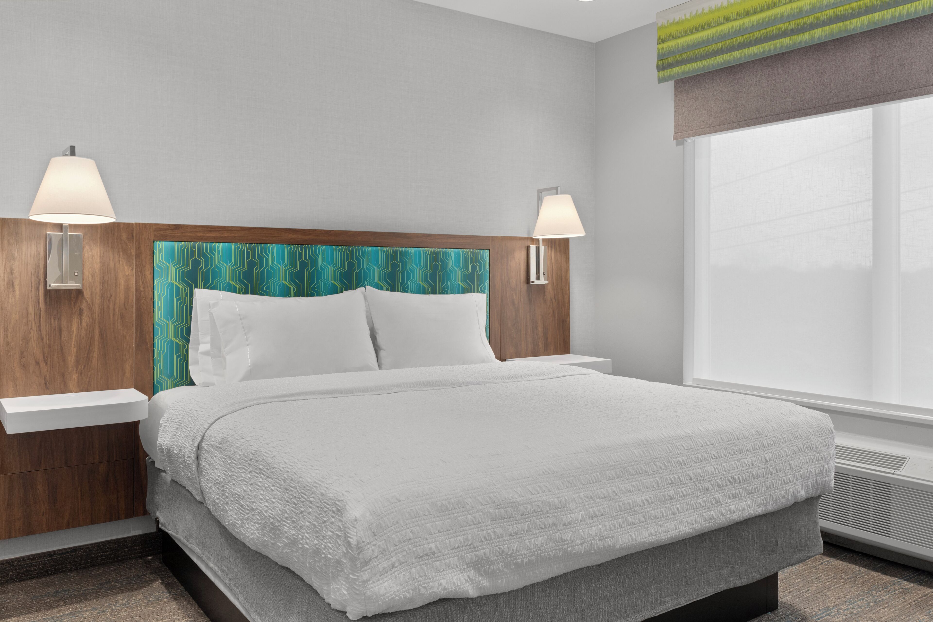 A neatly made bed with a white comforter and pillows, a wooden headboard with a teal pattern, two bedside lamps, and a large window with a view.