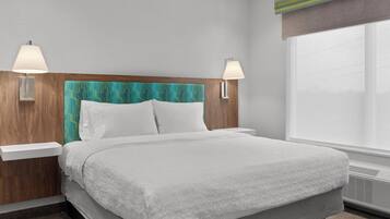 A neatly made bed with a white comforter and pillows, a wooden headboard with a teal pattern, two bedside lamps, and a large window with a view.