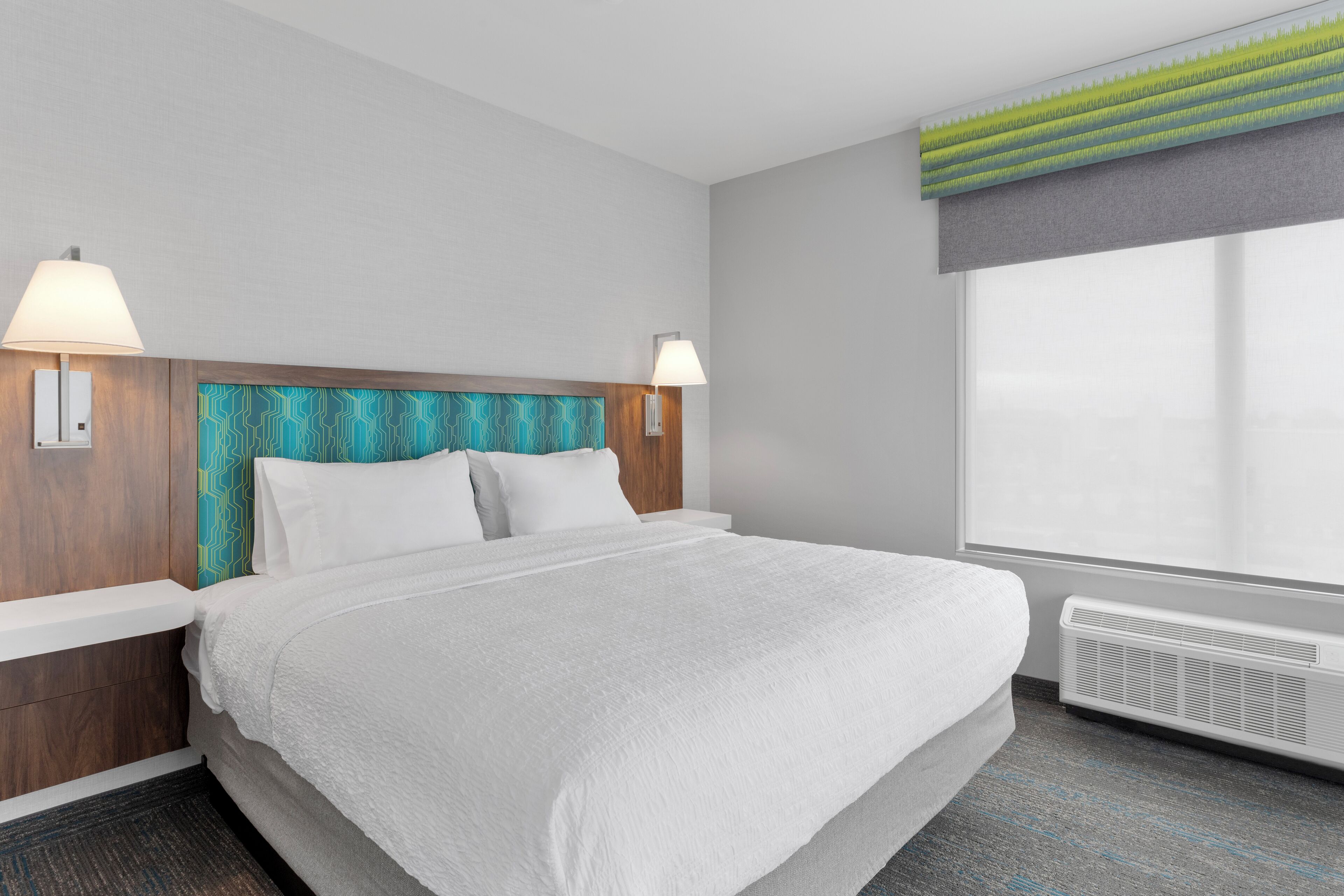 A hotel room with a large bed, two bedside tables with lamps, a headboard with a geometric pattern, a window with a view, and a wall-mounted air conditioner.