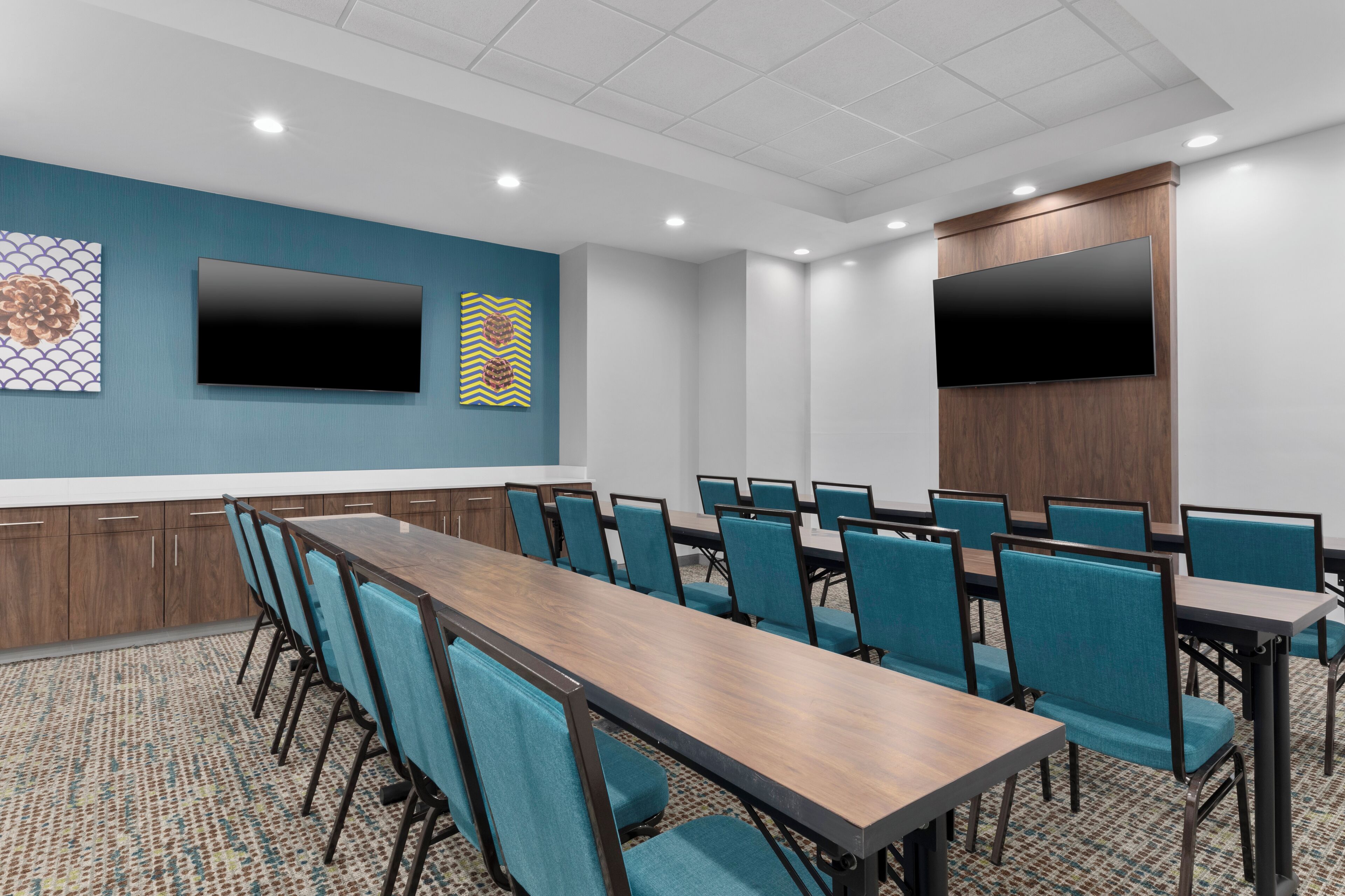 A conference room with a long table and chairs, two large flat-screen TVs mounted on the walls, and a patterned carpet.