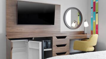 Une chambre d’hôtel moderne équipée d’un téléviseur à écran plat fixé sur un panneau en bois, d’un petit réfrigérateur avec un four à micro-ondes, d’une chaise jaune et d’une table de chevet avec une lampe.