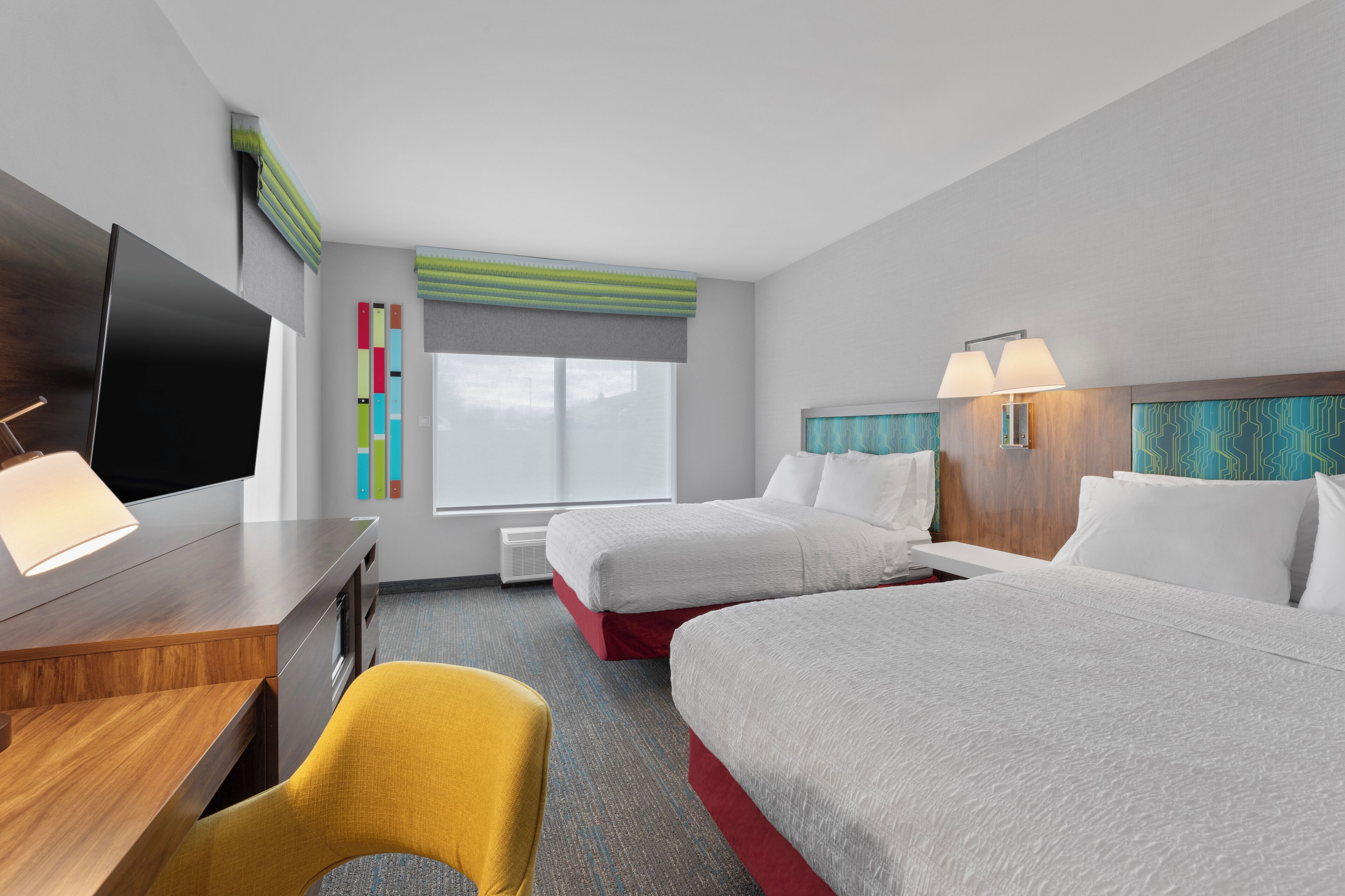 A hotel room with two beds, a desk with a TV, a chair, and a window with blinds.