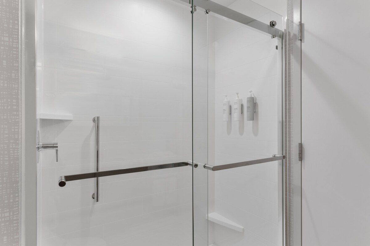 A modern shower with glass doors, a metal handrail, and a white tiled wall.